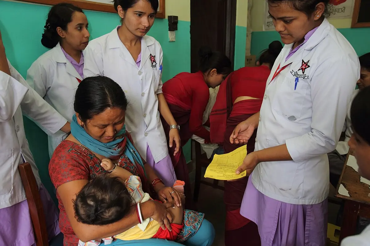 A health worker in a white coat gives an injection to a baby held on its mother’s lap, while other health workers stand nearby, one holding a yellow card
