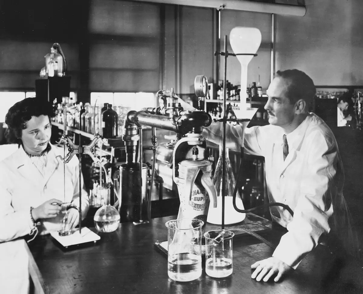 A black-and-white photo of a laboratory bench crowded with glass flasks and tubes; a young woman on the left works at the bench and a man in a white coat on the right leans on it