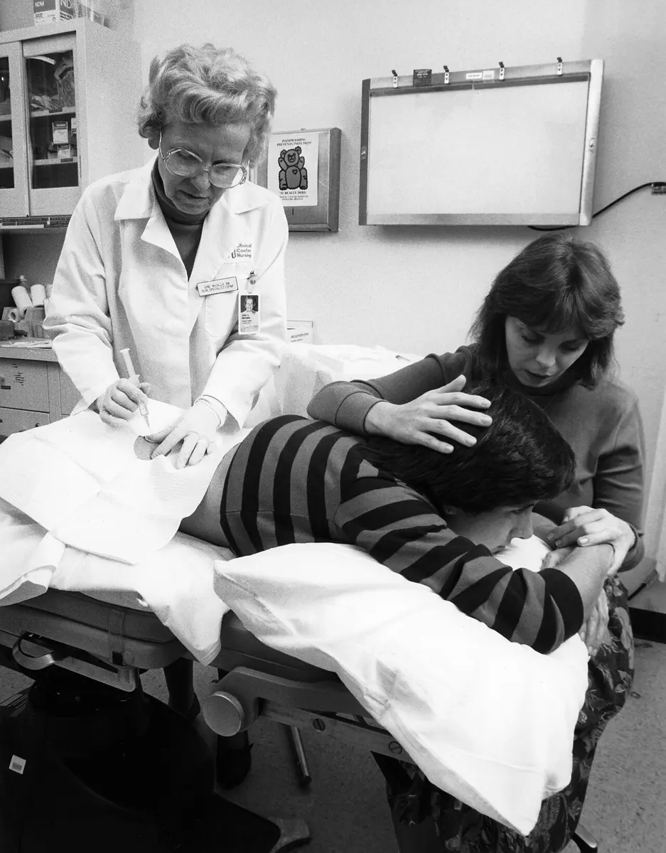 Black and white photo: a nurse in a white coat holds a syringe with a needle at the lower back of a teenage girl lying face down on an examination table, while a woman comforts the girl