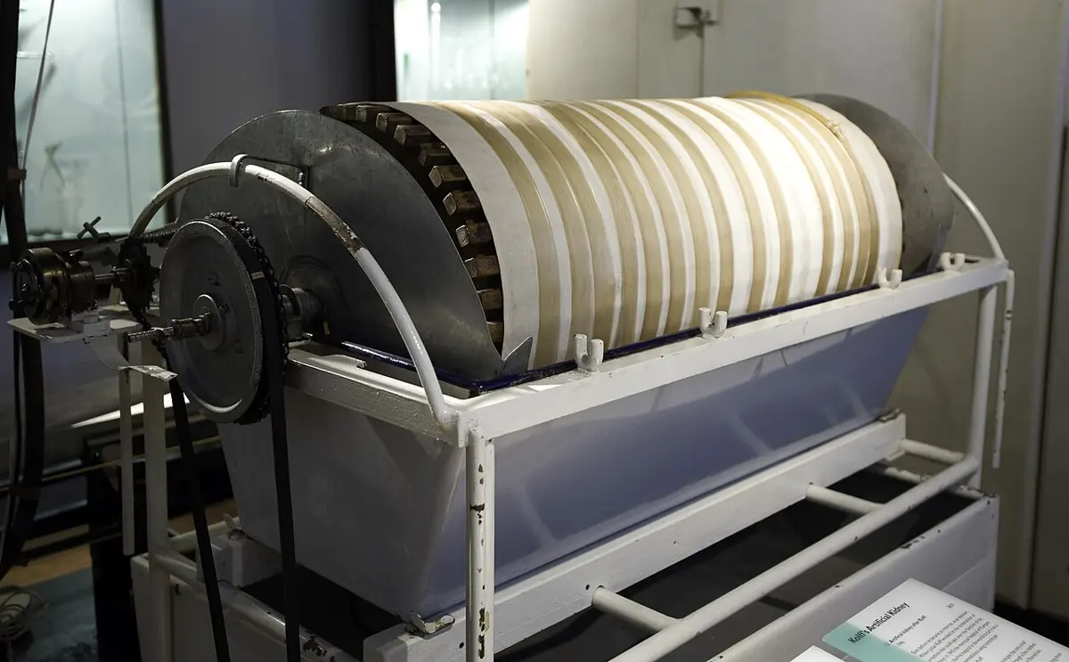 A large metal drum wound round with a long flat tube, sitting in a white trough on a metal frame, with a chain and gear to turn it, in a museum