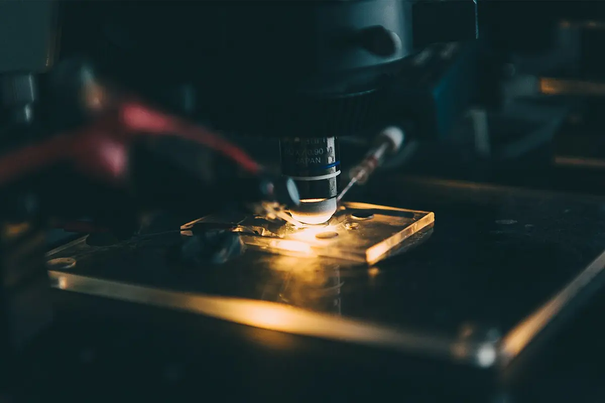 A microscope lens pointing down at a small glass dish lit brightly from below, with a thin glass pipette coming in at an angle from the right, in a dark room