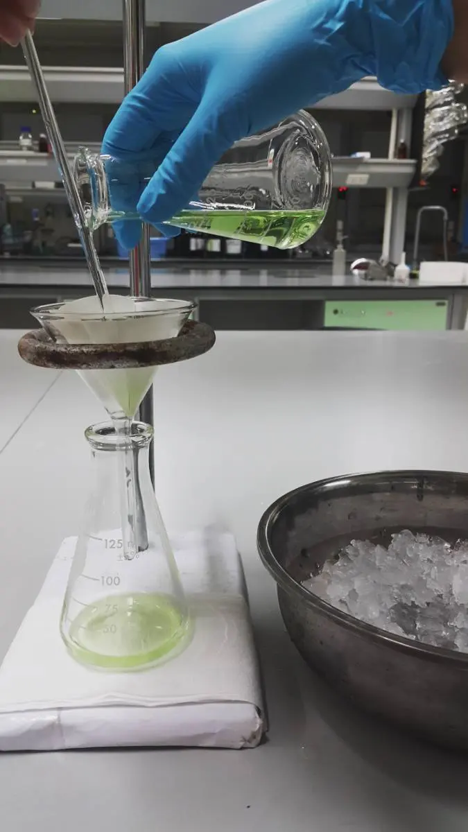 A gloved hand pours a pale green liquid from a beaker down a glass rod into a glass funnel lined with white filter paper. The funnel sits in a metal ring over a conical flask, which is collecting the clear liquid. A metal bowl of ice stands beside it.