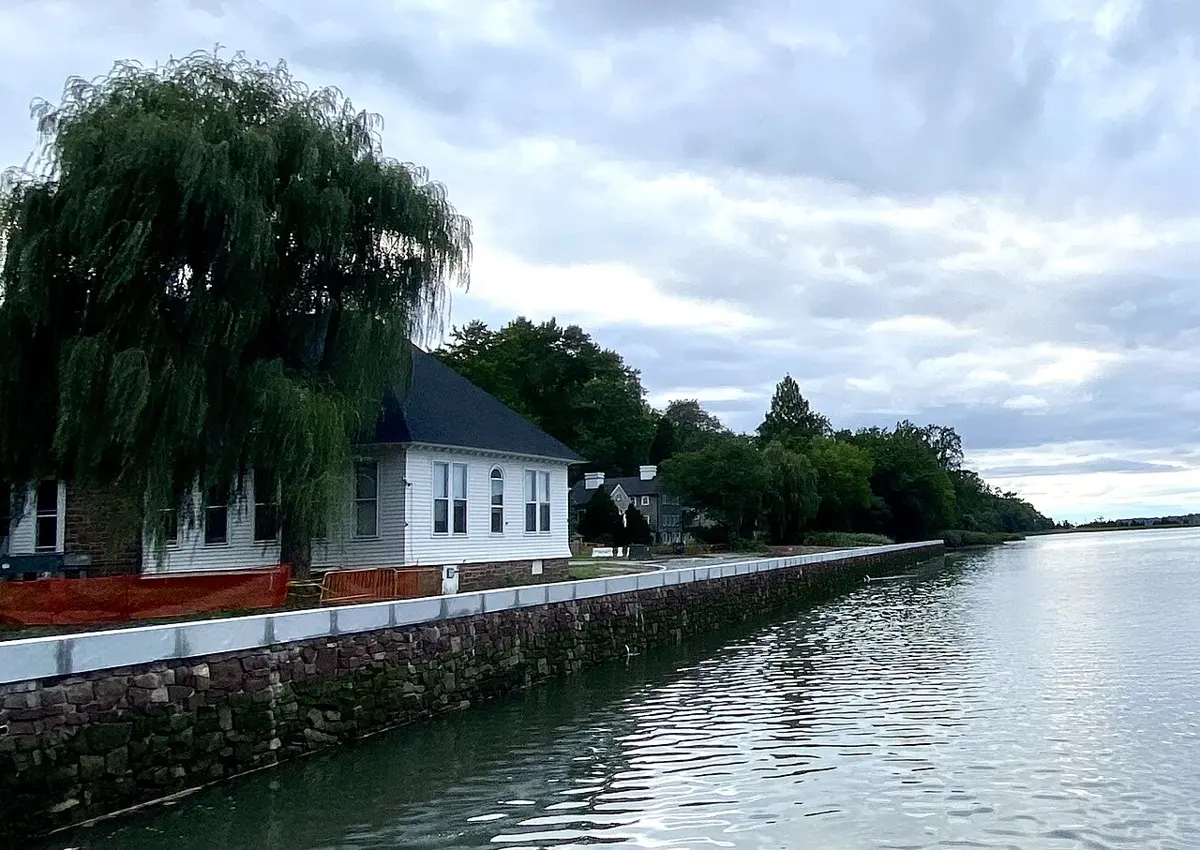 A white wooden house with a dark roof under a large willow tree, beside a calm stretch of water edged by a stone wall, with trees along the shore