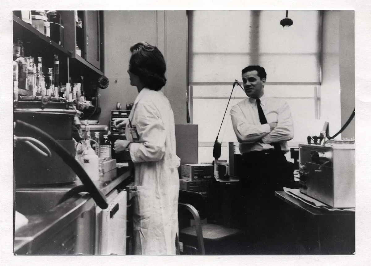 Black and white photo of a laboratory: a woman in a white coat works at a bench of bottles and equipment, while a man in a shirt and tie stands with arms folded, smiling