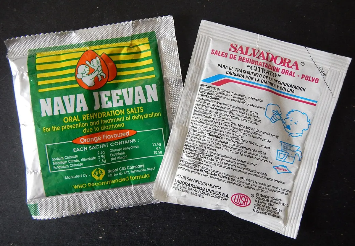 Two packets of oral rehydration salts on a dark surface: a green and silver Nava Jeevan packet from Nepal on the left, and a white Salvadora packet from Peru on the right