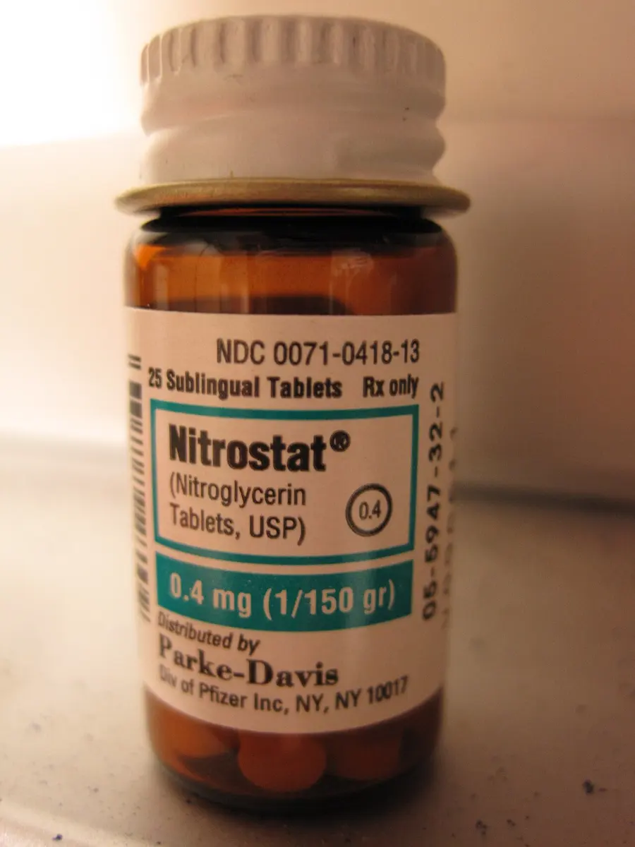 A small brown glass bottle with a white cap, labeled Nitrostat, Nitroglycerin Tablets, 25 sublingual tablets, 0.4 mg, with small round tablets visible at the bottom