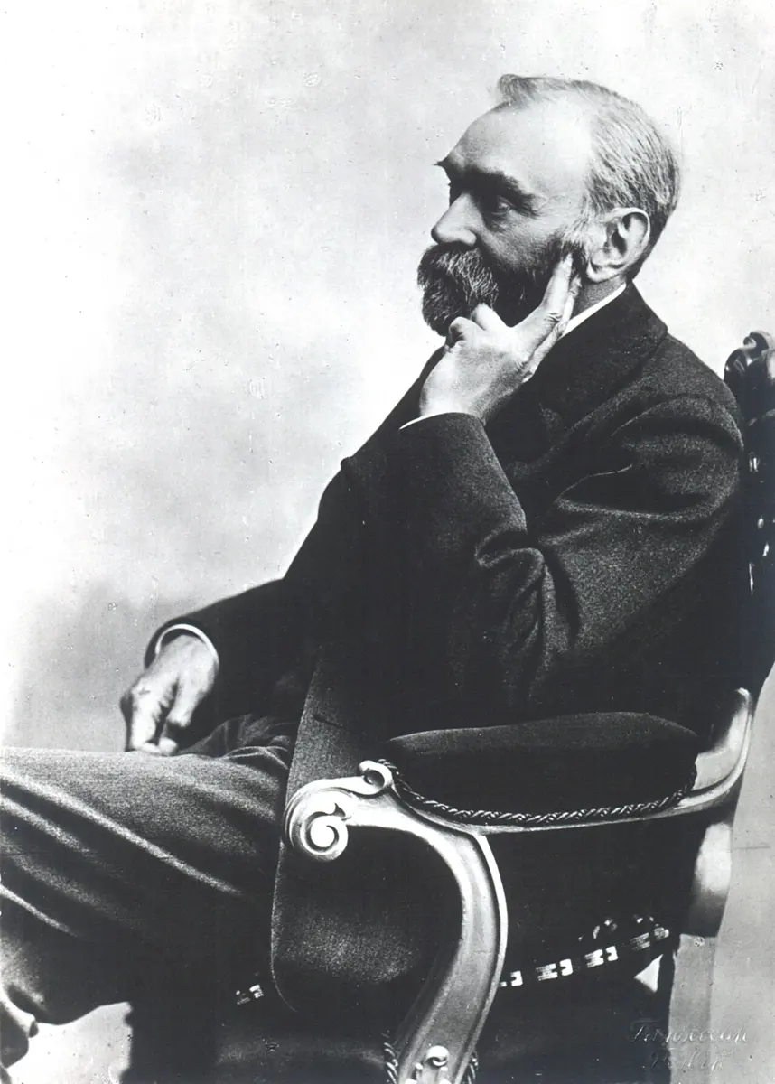 An old black-and-white photograph of a gray-bearded man in a dark suit, seen from the side, sitting in a carved armchair with one finger resting against his cheek