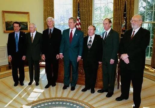 Seven men in dark suits standing in a row in a room with a round blue carpet and tall windows