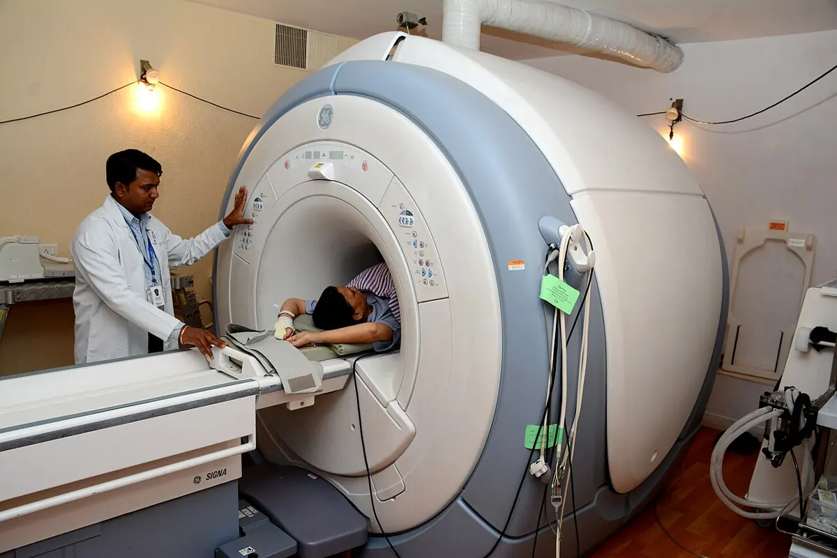 A doctor in a white coat stands beside a large white ring-shaped MRI scanner while a patient lies on the bed with her head inside the tunnel