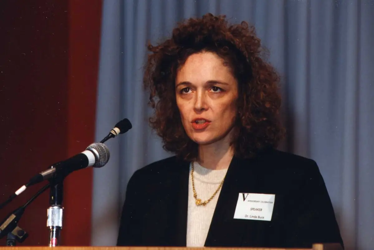 A woman with curly brown hair speaking into microphones at a lectern, wearing a dark jacket and a name badge
