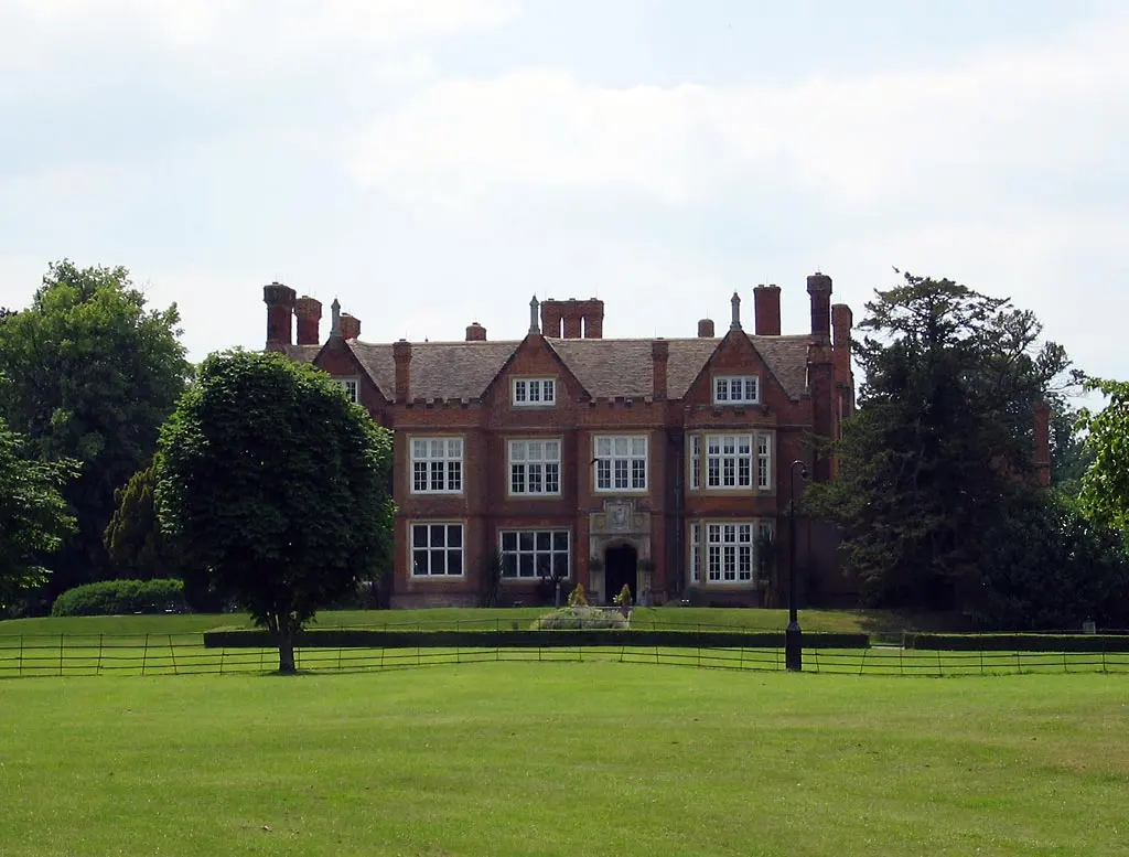 A large old red-brick house with tall chimneys and many white-framed windows, behind a wide green lawn and trees
