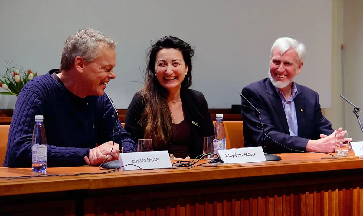 Edvard Moser, May-Britt Moser and John O'Keefe sitting side by side at a long table with name cards and microphones, smiling