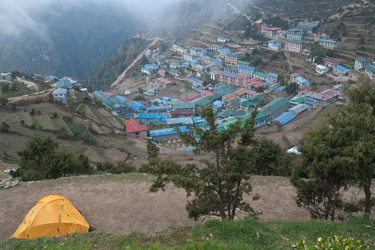 A horseshoe-shaped bowl on a steep mountainside filled with houses and lodges with blue, green and red roofs, a yellow tent on a grassy ledge in front, clouds above