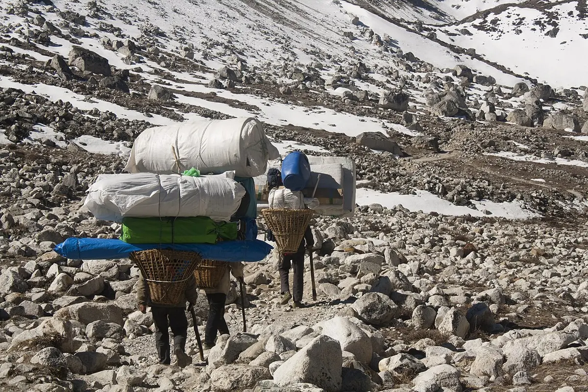 Two porters carrying tall loads on their backs walk up a rocky trail between snowy slopes