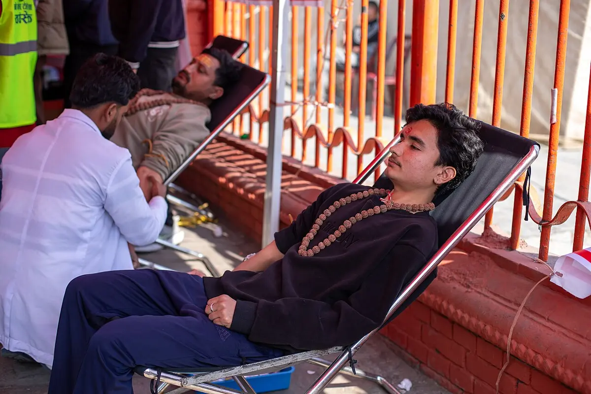 Two young men lying back on folding chairs beside a red-painted railing, giving blood, with a health worker in a white coat bending over one of them