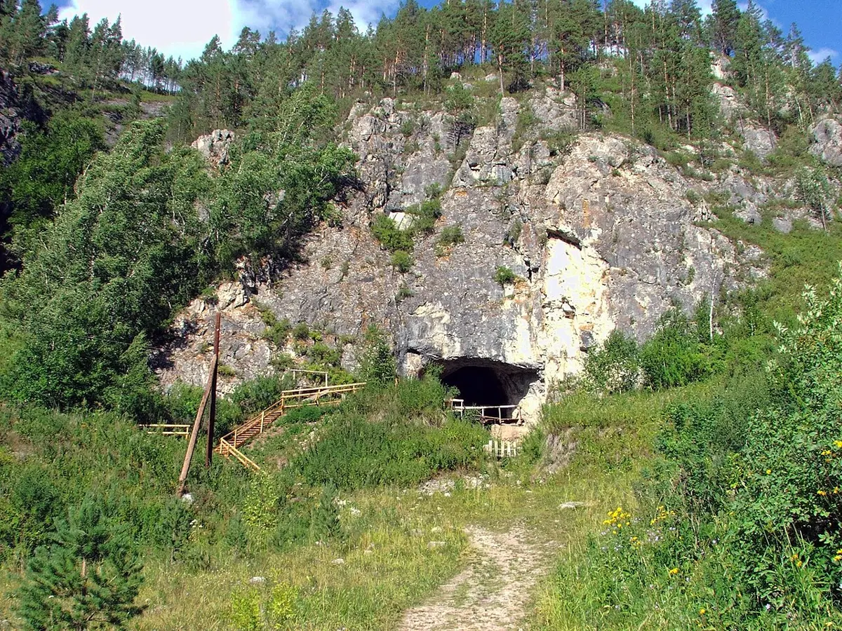 A low, dark cave mouth at the foot of a gray limestone cliff, with green trees on the slope above and a wooden walkway leading to the entrance