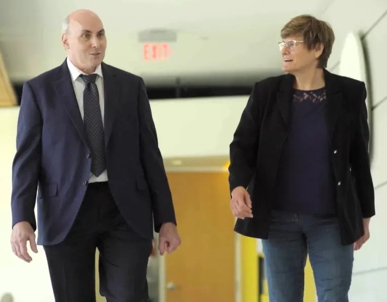 A man in a dark suit and tie and a woman with short brown hair and glasses walking side by side along a bright corridor, talking