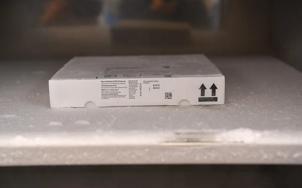A flat white cardboard box of vaccine vials, with a printed label and two upward arrows, lying on a frosty shelf inside a freezer