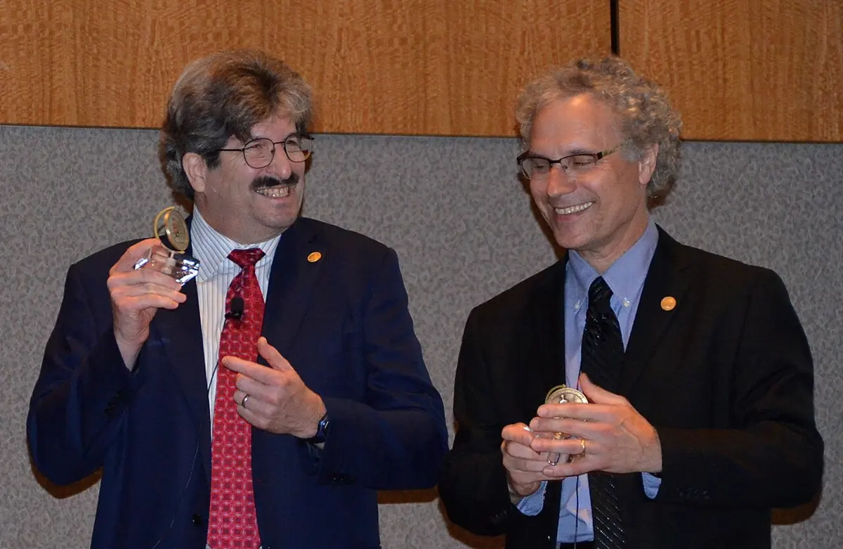 Two smiling men in suits holding glass prize awards; on the left Gary Ruvkun, with a mustache and a red tie, on the right Victor Ambros, with curly gray hair