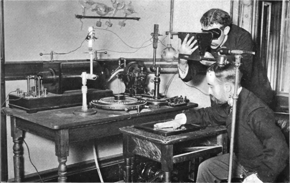 An old black and white photograph: a seated man rests his hand on a flat plate under a round glass tube on a stand; a second man holds a box-shaped viewer to his eyes and looks through it at his own hand held in front of the tube; electrical apparatus fills the table behind them