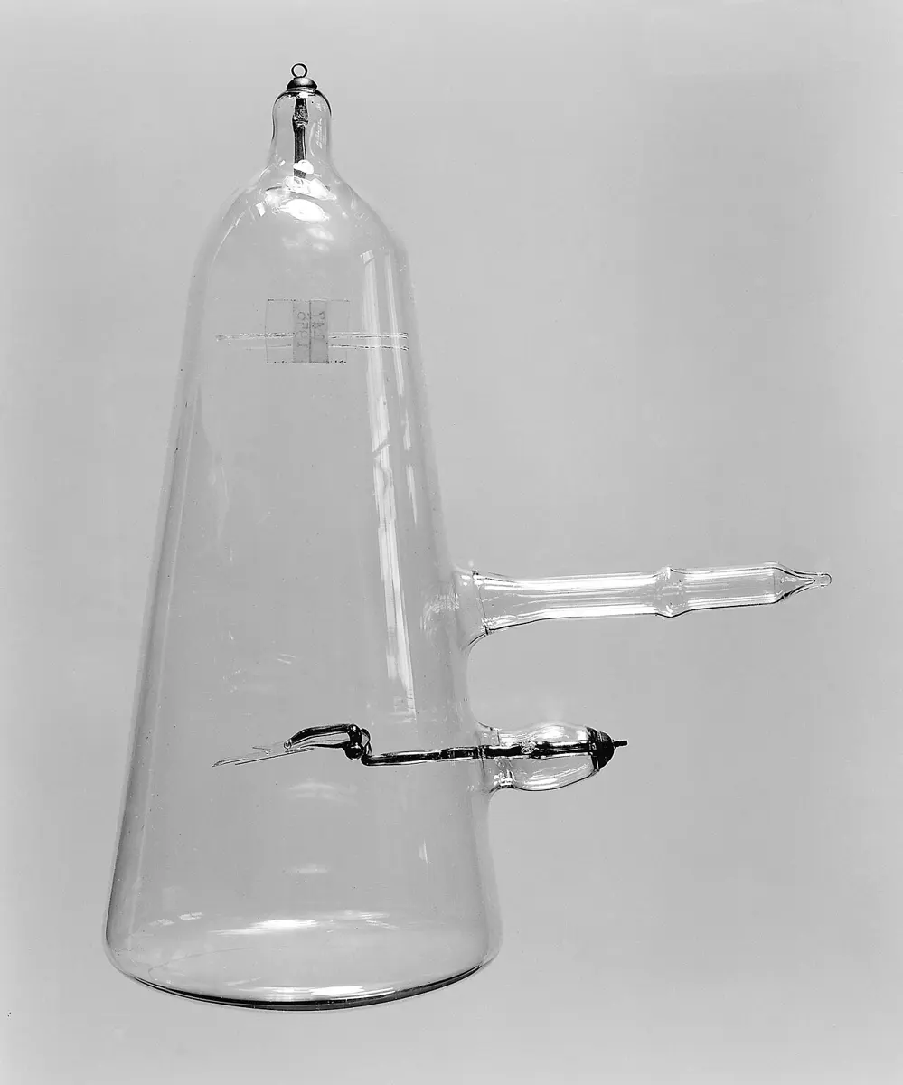 A tall, cone-shaped clear glass tube hanging from a metal ring at its narrow top, with a metal wire sealed in at the top, a glass side arm, and a metal cross on a hinged rod low down, seen edge-on