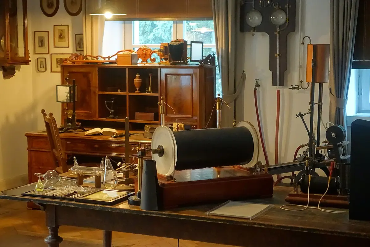 An old laboratory: on a long wooden table a large induction coil, a drum wrapped in black, with glass discharge tubes and a framed plate beside it; behind, a wooden cabinet, framed portraits and a window