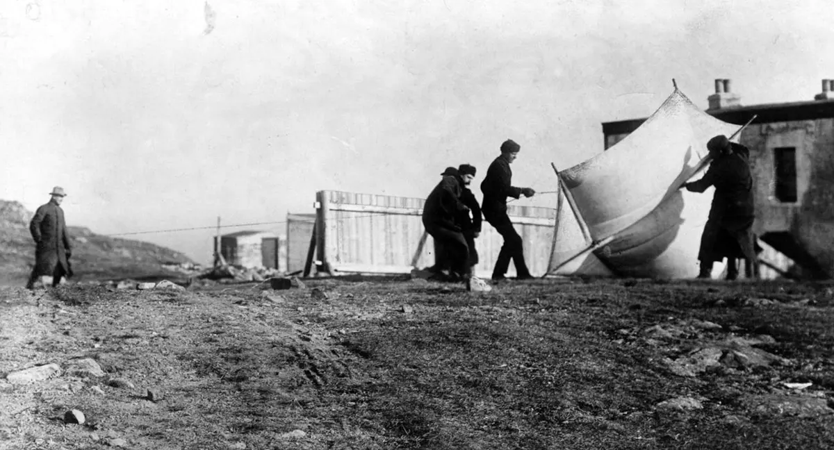 An old black-and-white photo of a bare, rocky hilltop. Four men in heavy coats are struggling to hold up a large white kite in the wind, beside a wooden hut and a low building.
