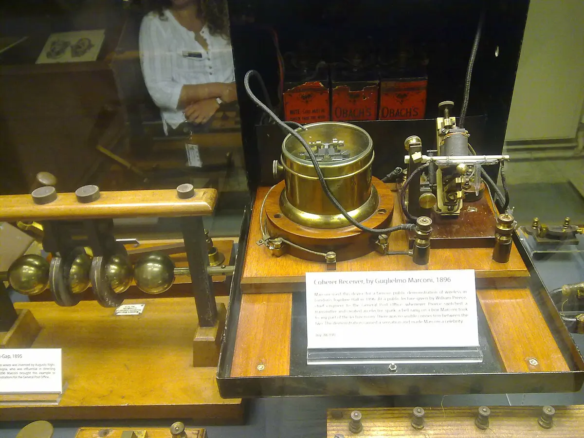 Old brass and wooden equipment in a museum glass case: a set of brass balls on a stand on the left, and on the right a wooden base with a round brass bell, a small glass tube held between clips, and a printed museum card