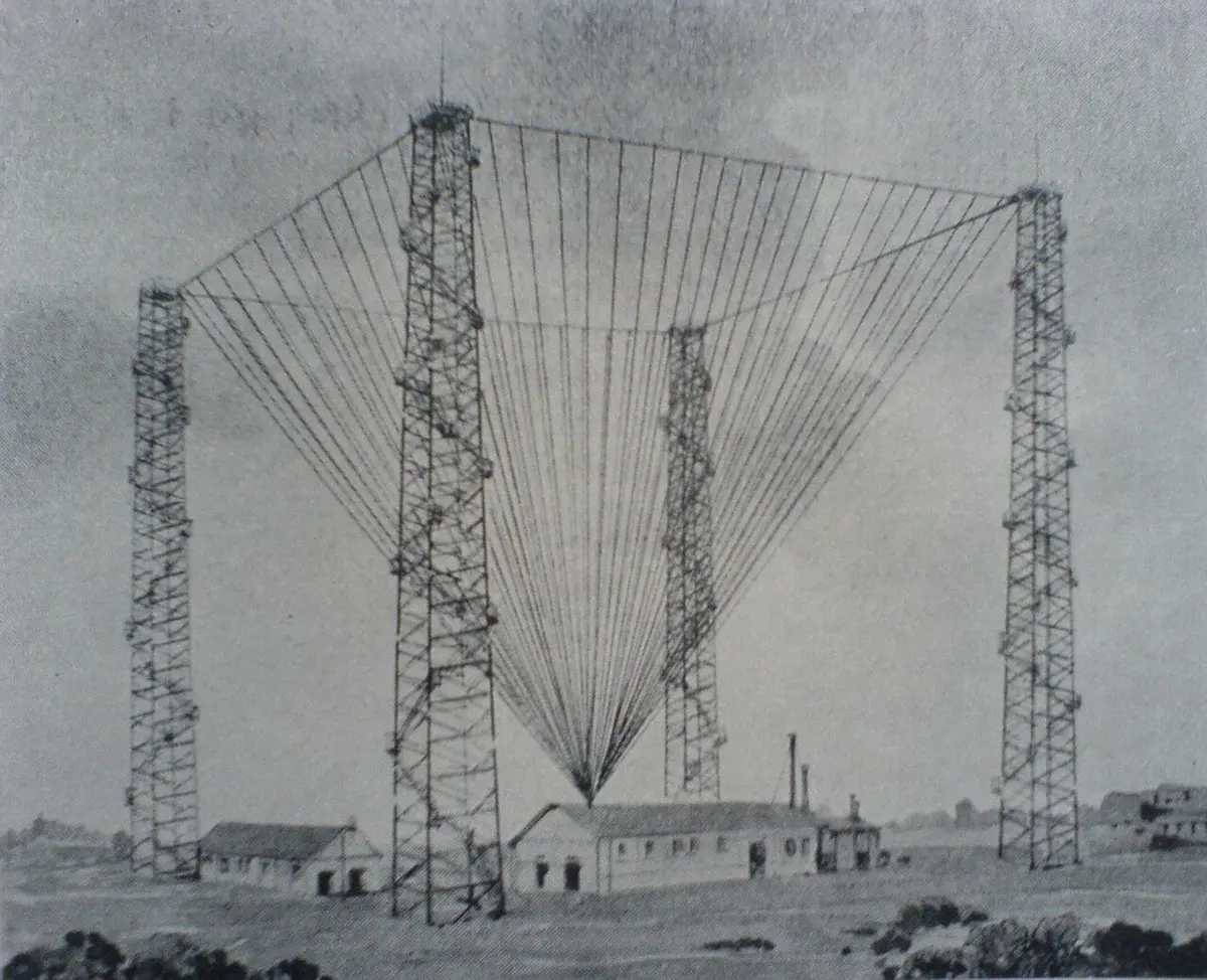 An old drawing of a wireless station by the sea: four tall wooden lattice towers at the corners of a square, with dozens of wires hanging from a rope between them and gathering in a cone down into a low building