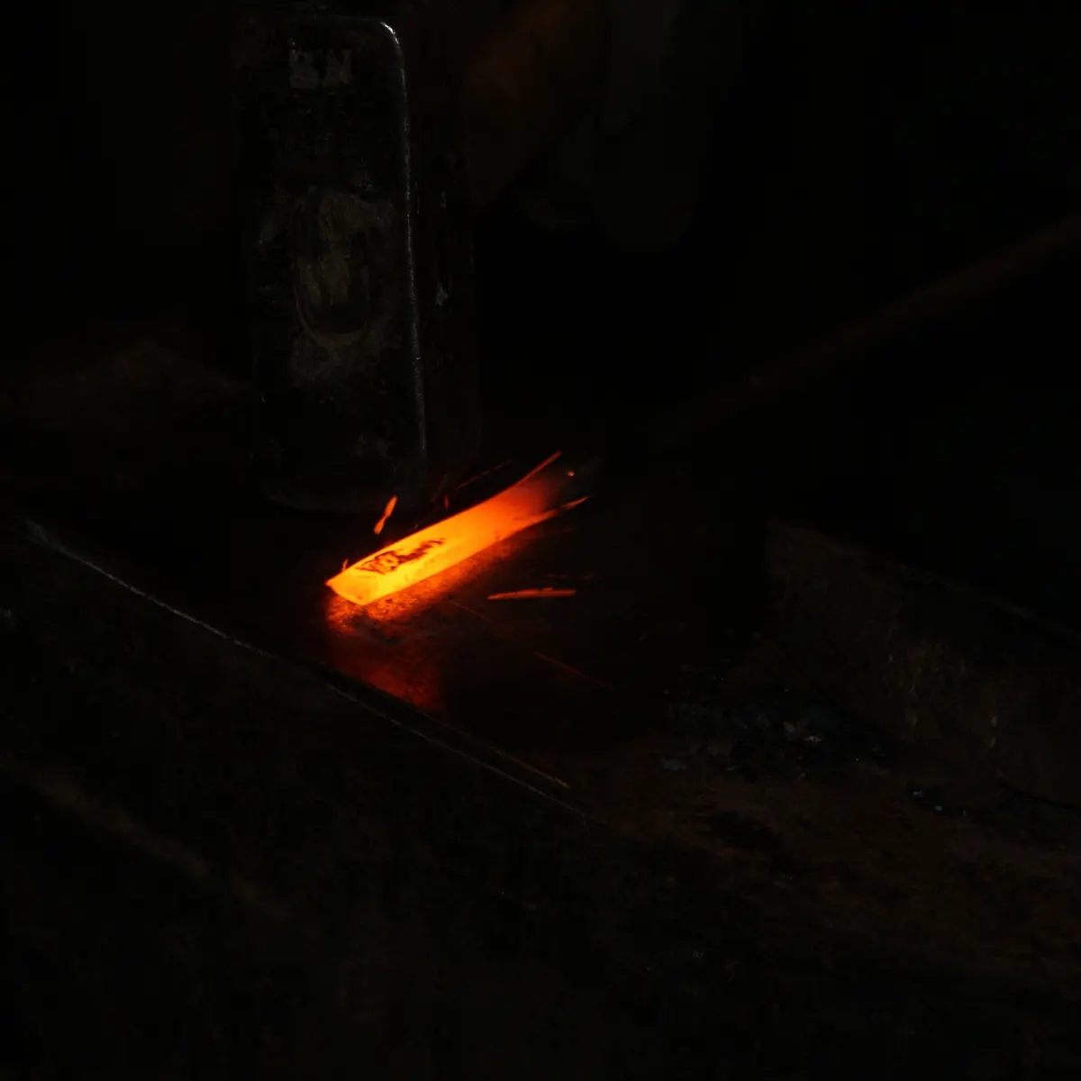 In a dark workshop, a short iron bar glowing bright orange lies on an anvil under the head of a hammer, with a few sparks flying