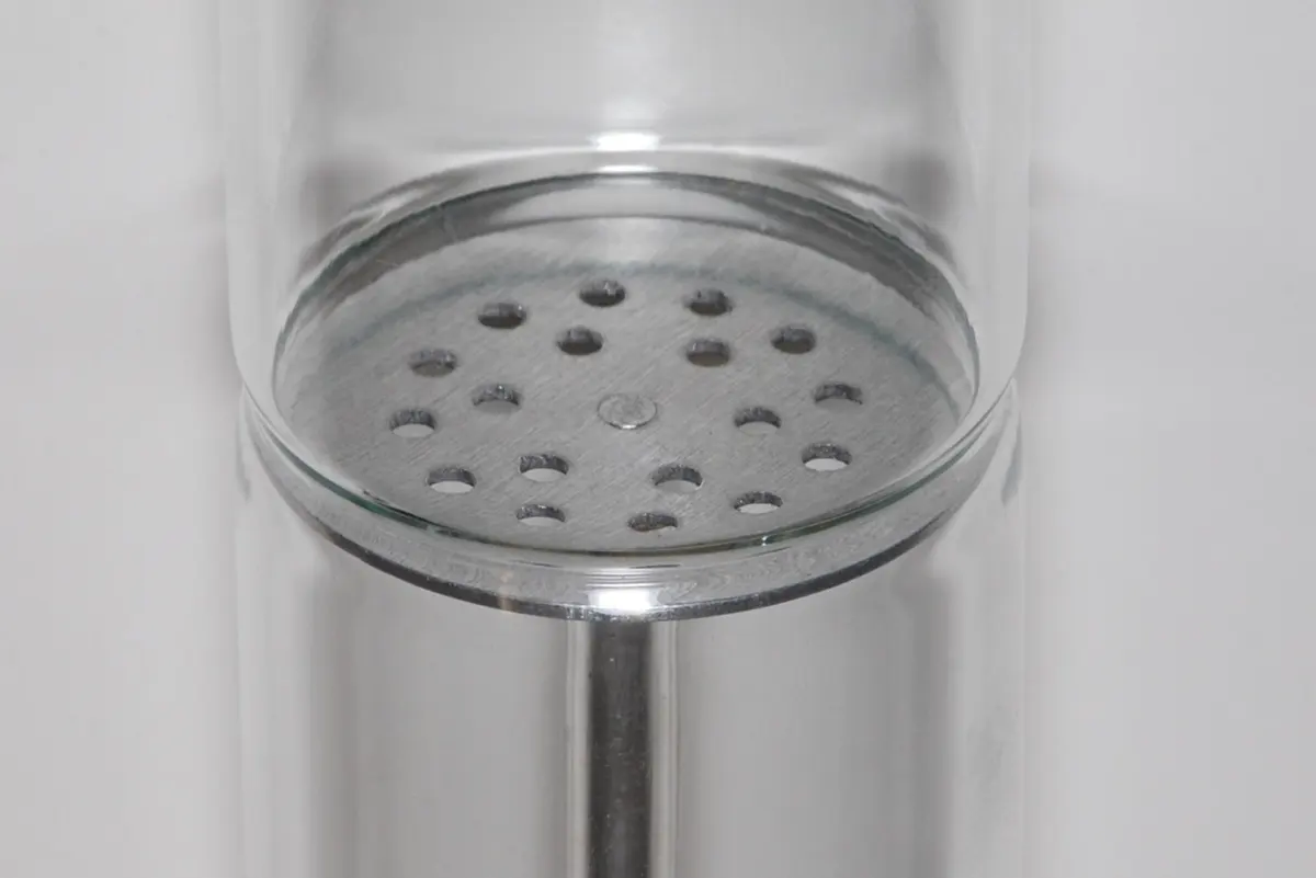 Close-up of a round gray metal disc fixed across the inside of a glass tube, with about twenty small round holes in it