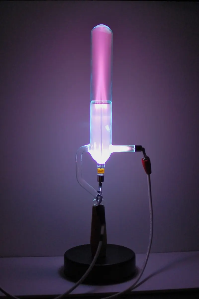 An upright glass tube on a stand, glowing pink at the top and white-blue lower down, with a side arm joined to a red electrical lead