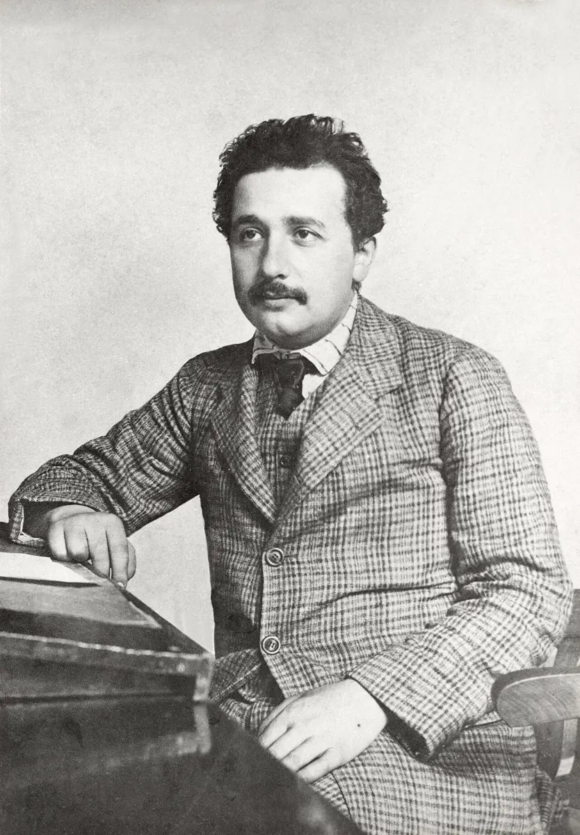 Albert Einstein as a young man, with curly dark hair and a mustache, in a checked suit, sitting with one arm resting on a sloping wooden desk