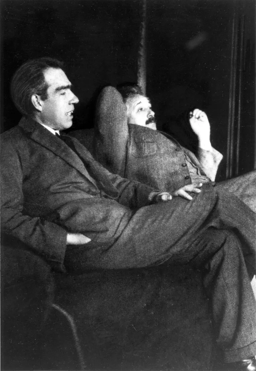 An old black and white photograph of two men in suits talking in armchairs: Bohr, in front, looks to the right and speaks; Einstein leans back behind him, smiling, one hand raised
