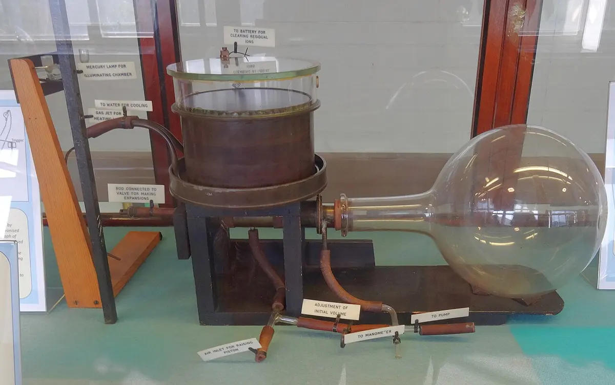An old apparatus in a museum case: a short brass cylinder with a glass top on a wooden stand, connected by pipes to a large round glass flask, with small labels on its parts