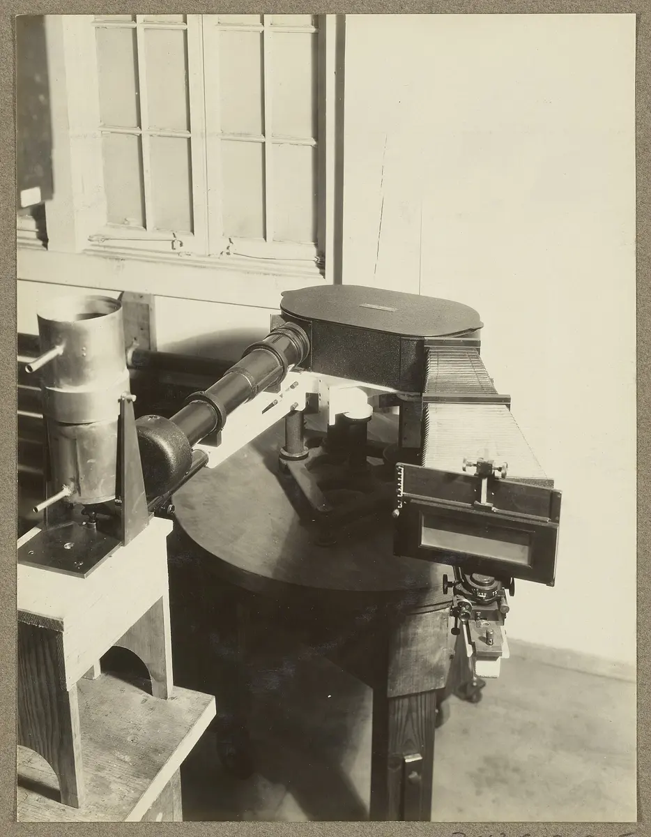 An old sepia photograph of laboratory apparatus on a round wooden table: a metal tube with water pipes holding a lamp, on the left, aimed through a long black tube into a large dark spectrograph box with a plate holder on its far end