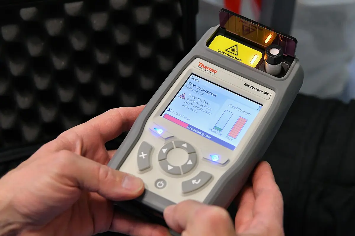 Two hands holding a gray handheld instrument with buttons and a screen reading Scan in progress; at its top, a small glass bottle sits in front of a yellow panel marked Laser Aperture