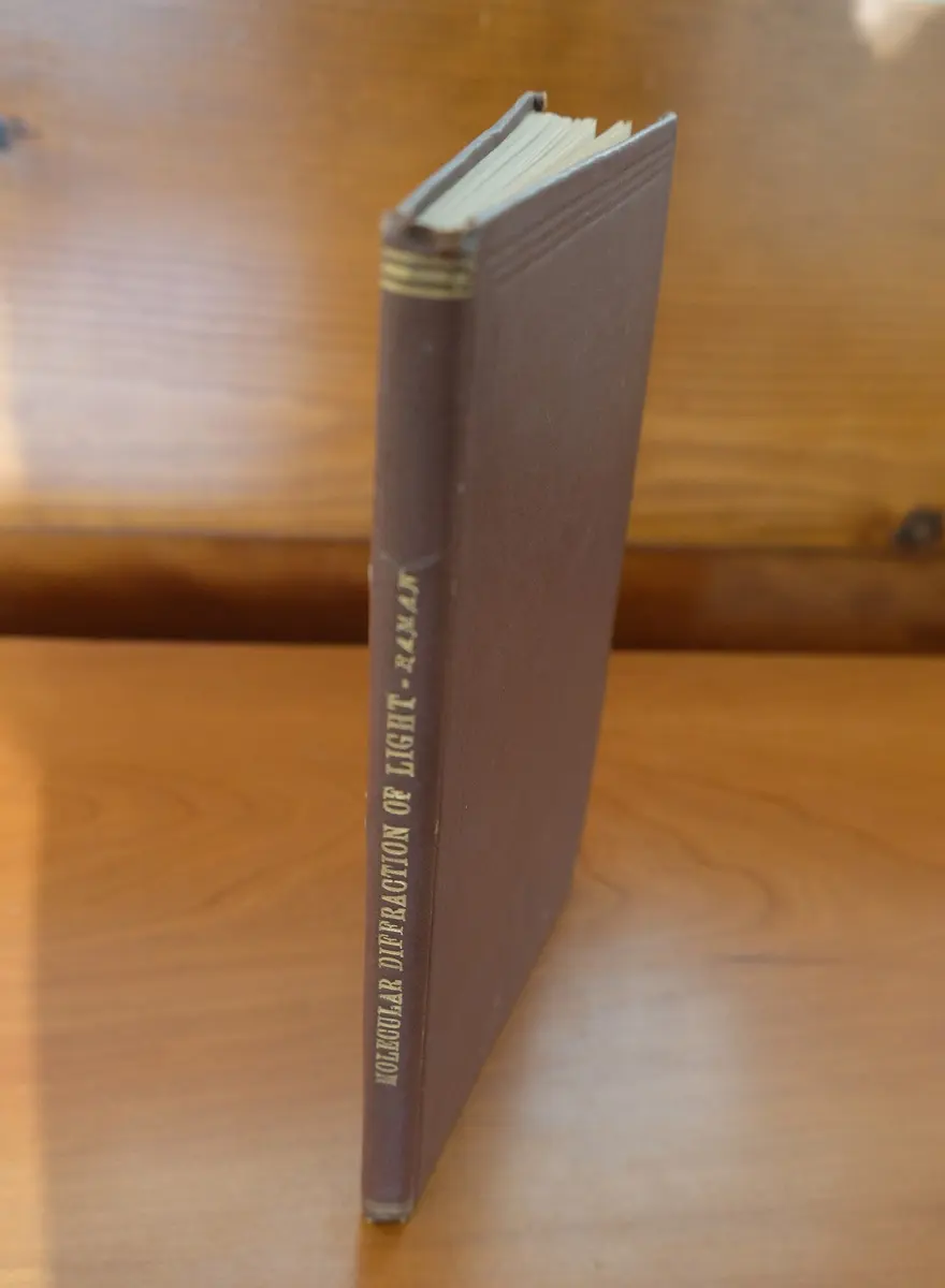 A slim old hardback book in a brown cloth cover standing on a wooden table, its spine lettered in gold: Molecular Diffraction of Light, Raman