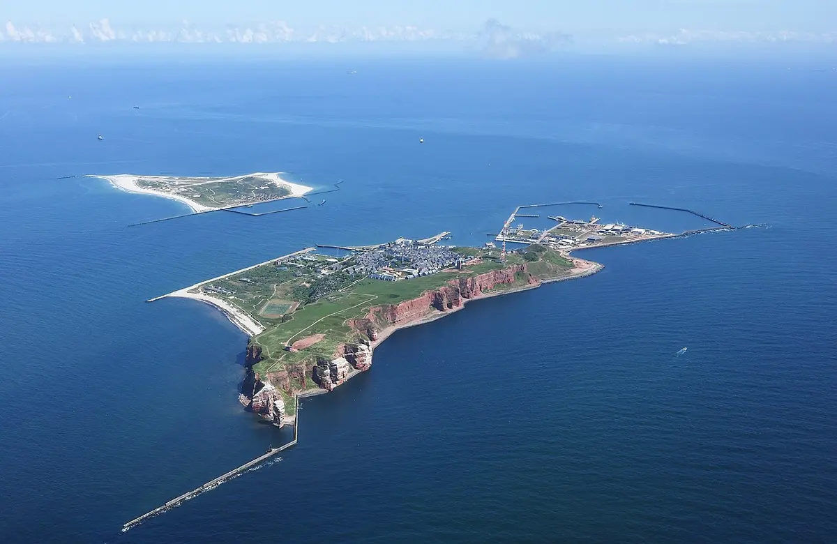 An aerial photo of a small island in a deep blue sea, with red cliffs, a harbor and a town, and a smaller sandy island beside it