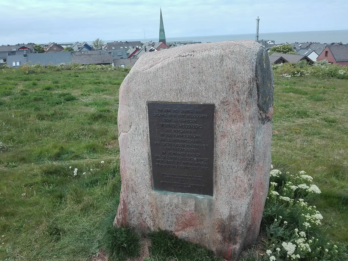 A large pinkish boulder standing on grass, with a dark metal plaque fixed to it; houses and a church spire behind