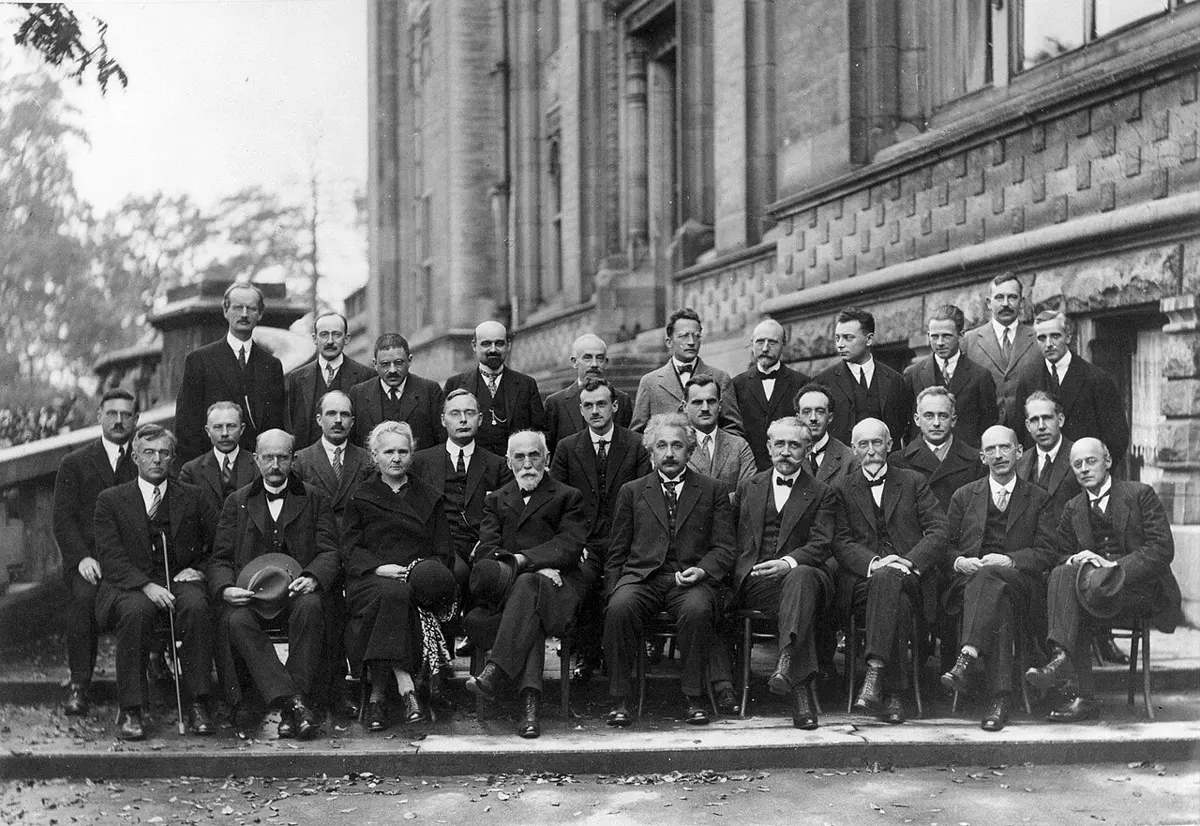 An old black-and-white group photo of twenty-nine scientists in dark suits, all men except one woman, in three rows in front of a building