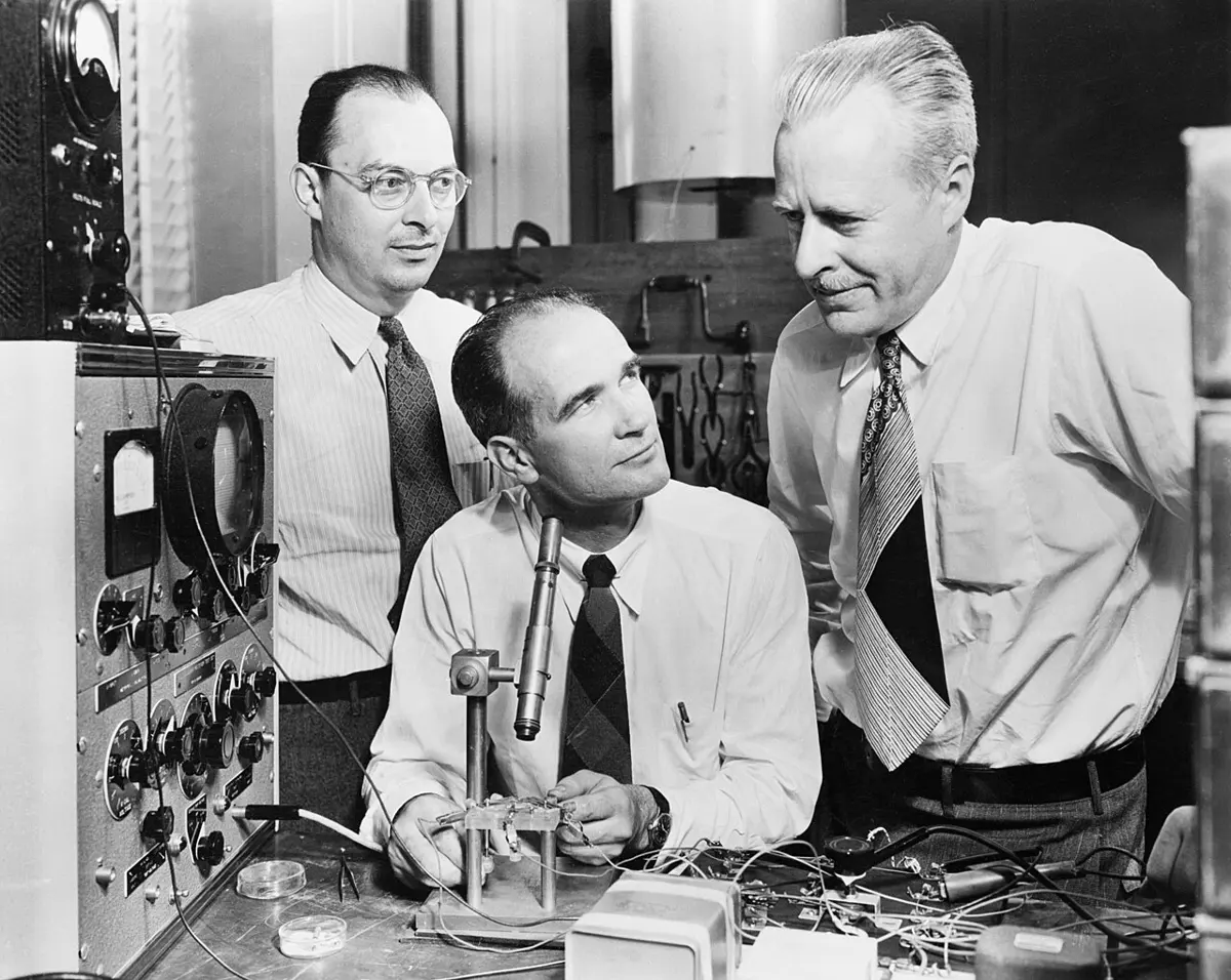An old black and white photograph of three men in shirts and ties in a laboratory: one in glasses stands on the left, one sits at a bench with a small apparatus and a microscope in front of him, and an older man stands on the right, all looking at the apparatus