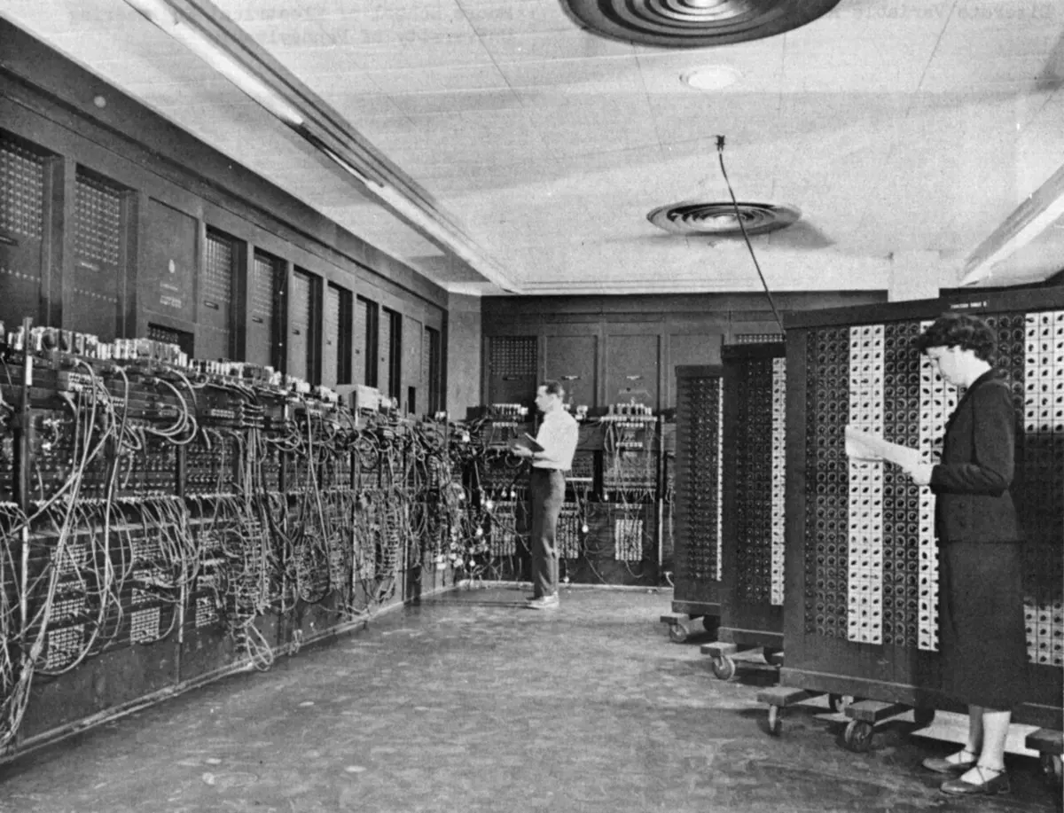 An old black and white photograph of a long room lined with tall dark panels covered in sockets, knobs and tangles of cables; a man stands at the far end and a woman in front reads a sheet of paper beside a panel