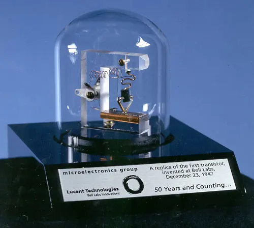 A small apparatus under a glass dome on a black base: a clear plastic block holding a coiled spring and a small triangle pressed down onto a copper-colored strip; a plaque reads A replica of the first transistor, invented at Bell Labs, December 23, 1947