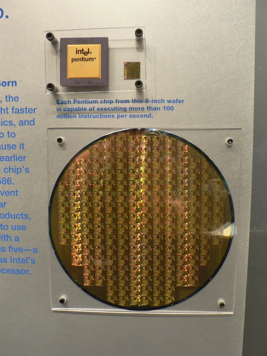 A round, shiny gold-colored silicon wafer covered in a grid of small square chips, on display in a museum; above it, a packaged Intel Pentium chip and a single bare chip