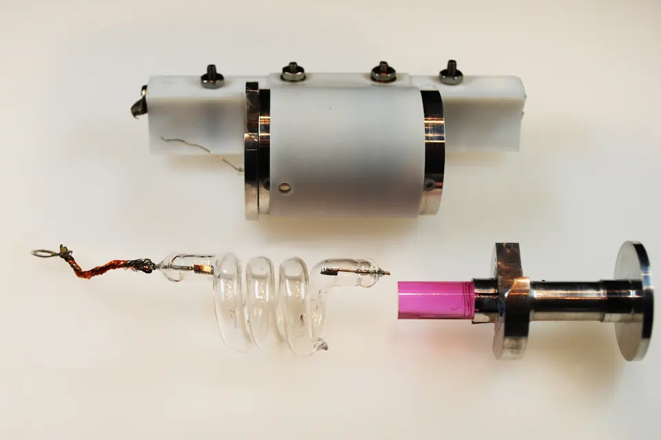 Three parts on a white table: a white cylinder with metal rings and screws, a clear glass tube coiled in a spiral, and a short pink ruby rod fixed to a shiny metal holder