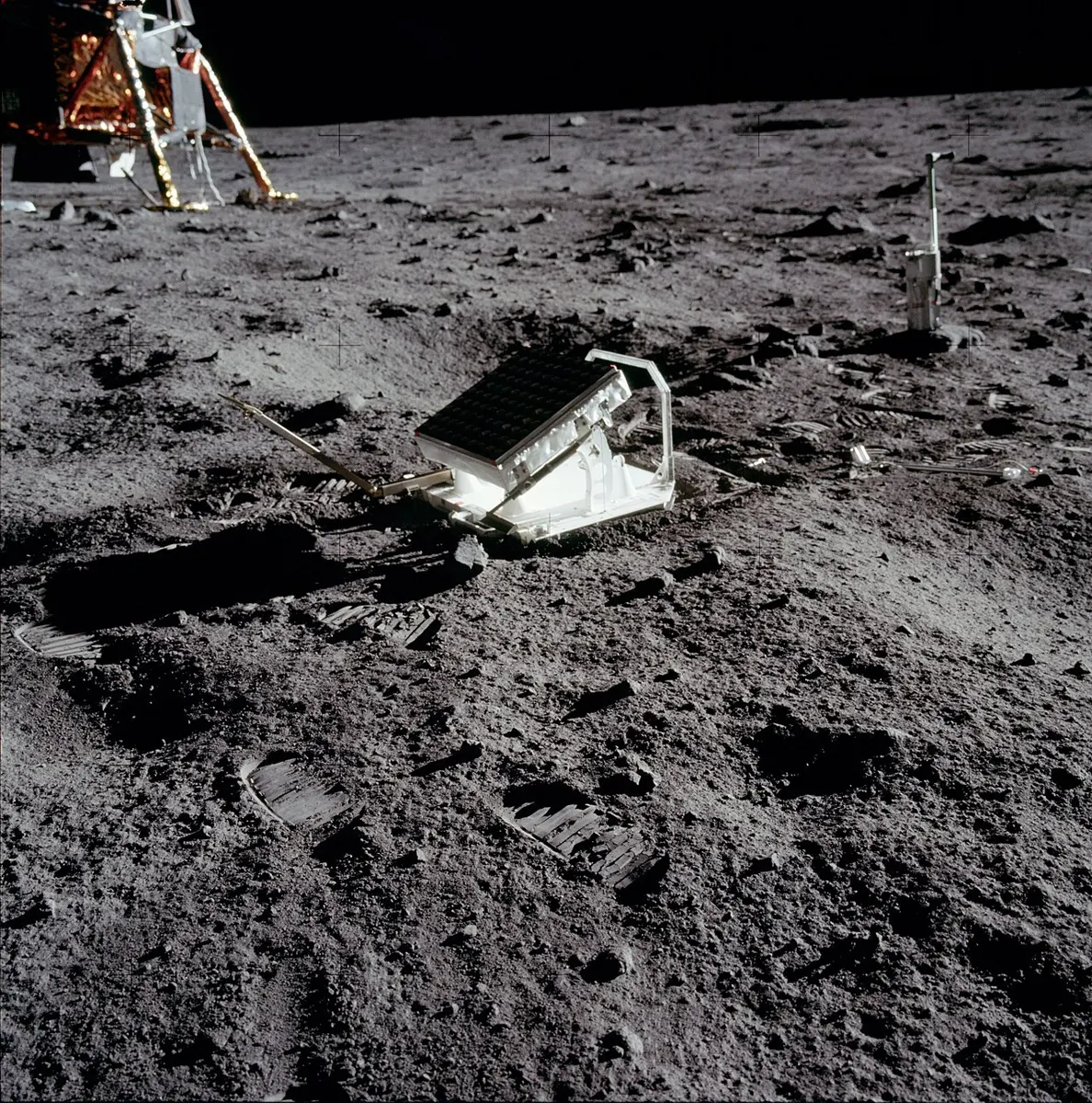 A flat panel of small round mirrors on a tilted metal stand on the gray, dusty ground of the Moon, with boot prints around it and the leg of the lunar module in the corner