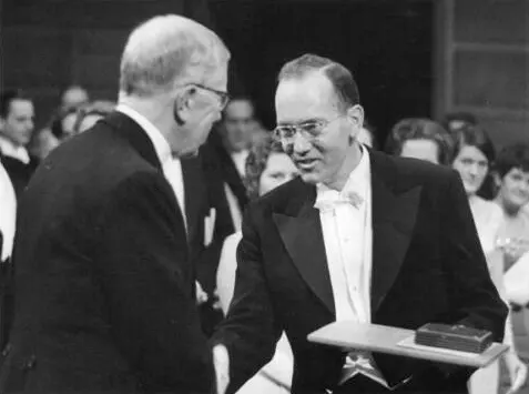 A black and white photograph: a smiling man in evening dress, holding a flat case, shakes hands with an older man seen from behind, in front of a seated audience