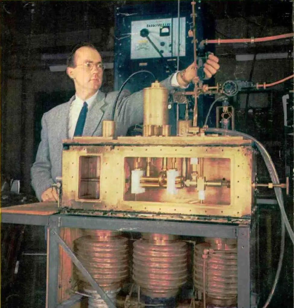 An old color photograph: a man in glasses and a gray jacket adjusts a valve on a long brass box with windows, full of pipes and fittings, standing on a frame above three big round pumps