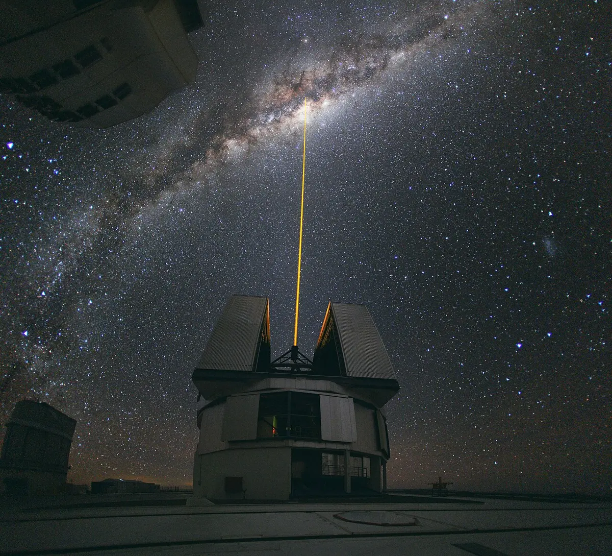 At night, a straight yellow-orange laser beam shoots up from an open telescope dome into a sky full of stars, towards the bright band of the Milky Way